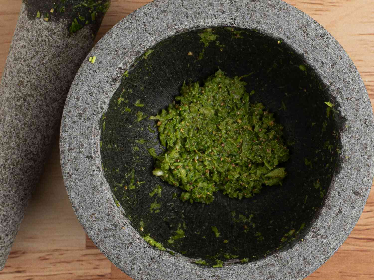 Garlic, cilantro stems, and peppercorns pound into a fine paste inside mortar, with pestle on the lefthand side of the image.