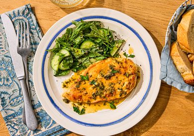 A plate with chicken paillard and a side of greens, placed on a wooden table with a fork and a napkin
