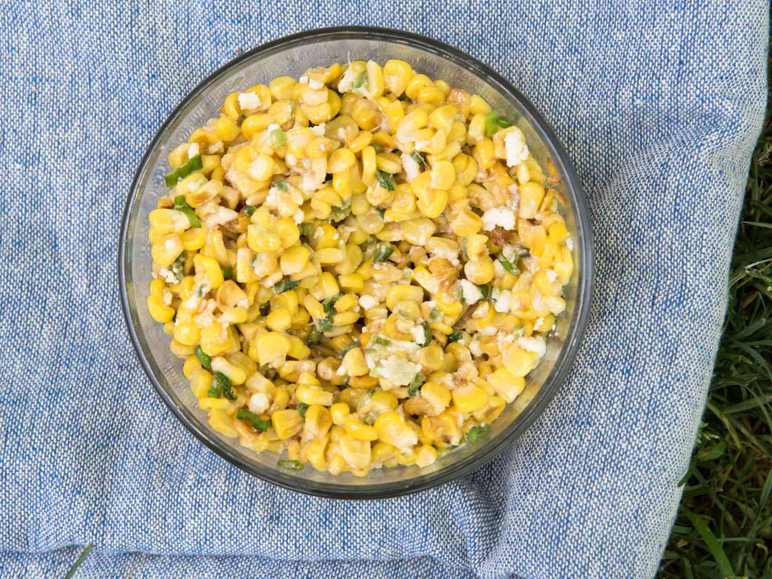 A glass bowl of esquites (corn salad) on a chambray blue cloth on grass.