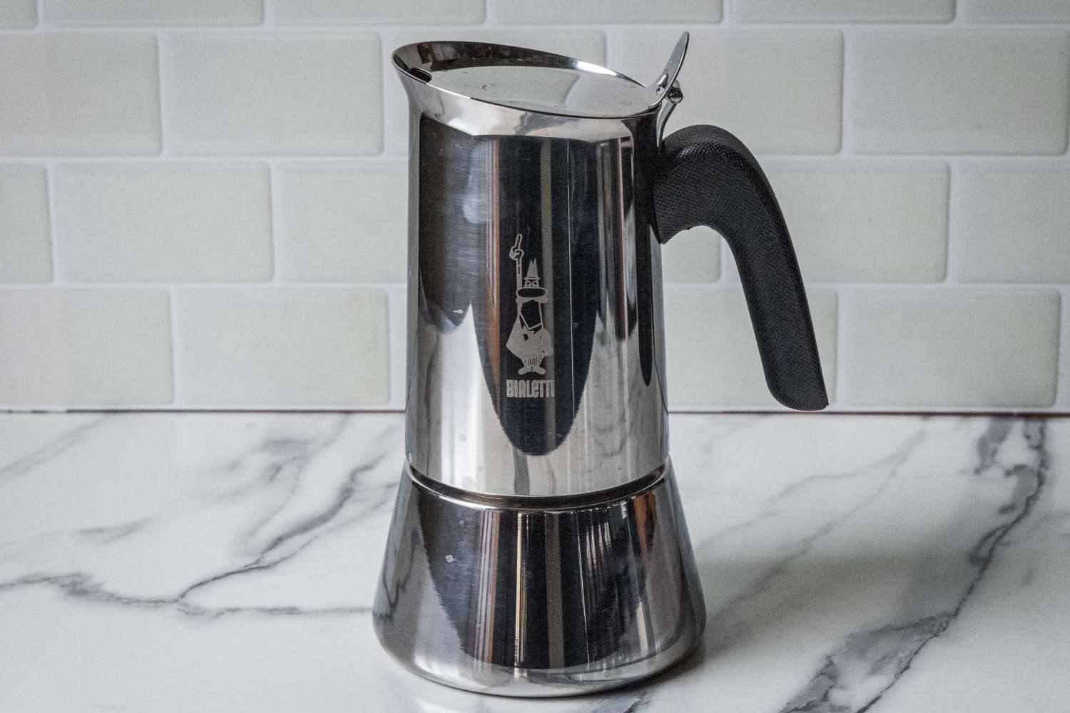 A stainless steel stovetop espresso maker with a black handle set on a marble surface