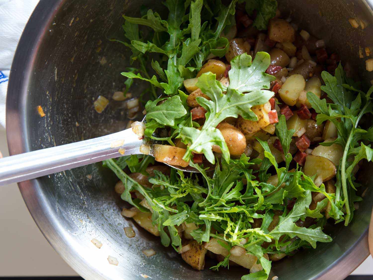 Mixing arugula leaves into a bowl of fingerling potatoes, sautéed onion, pickled onion, and diced chorizo.