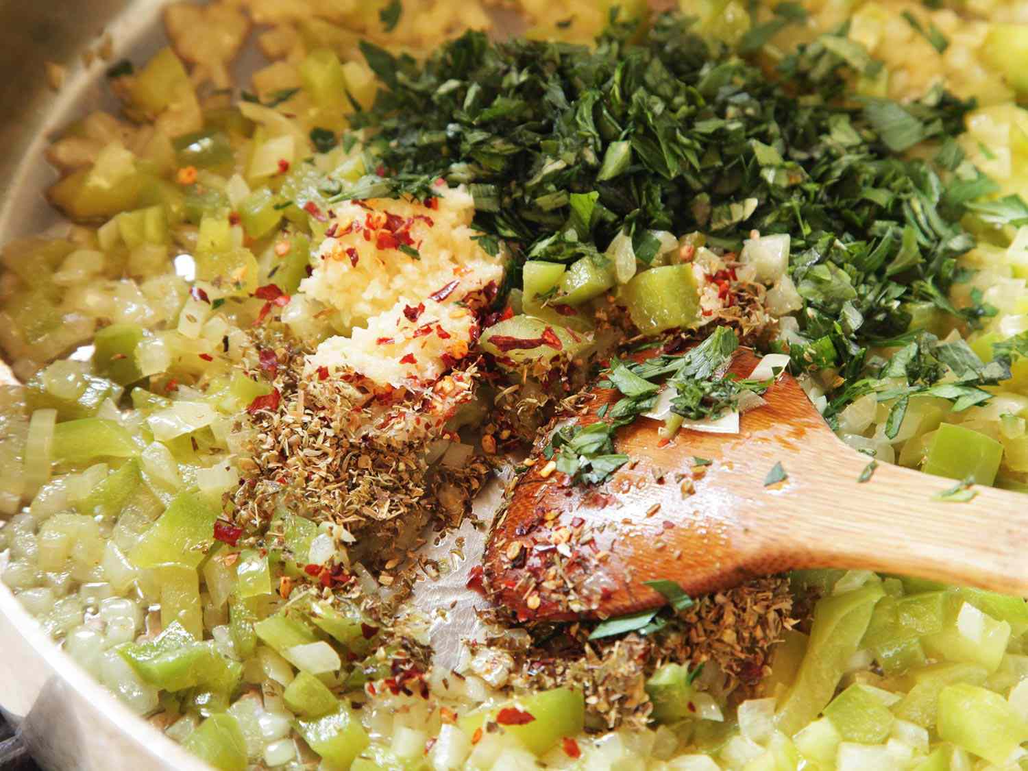 Close-up of galric, oregano, chile flakes, and parsley being stirred into the onions and green peppers with a wooden spoon.