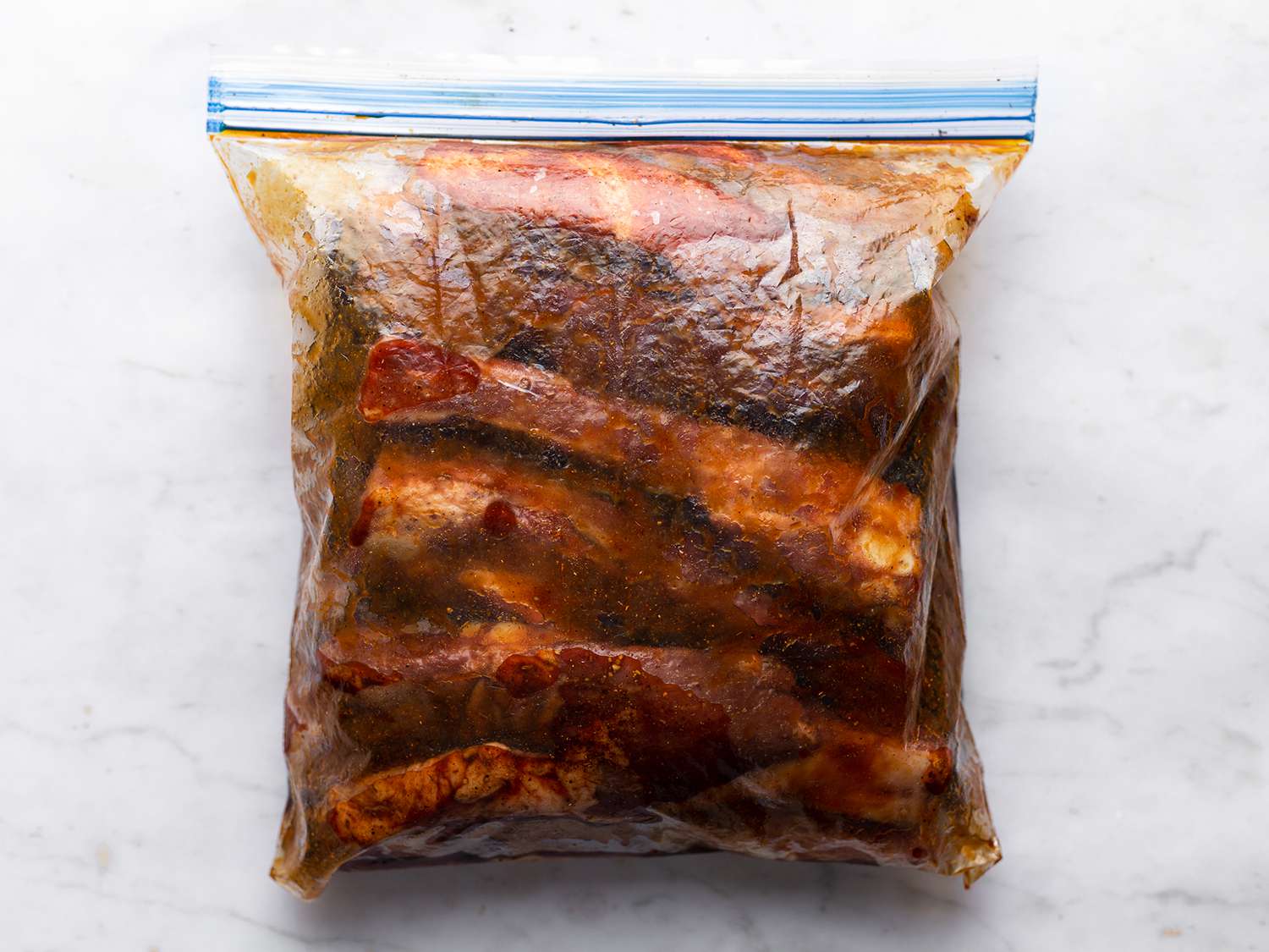 Ribs and liquid marinade inside of a sealed, zipper-lock bag, on a white stone background.