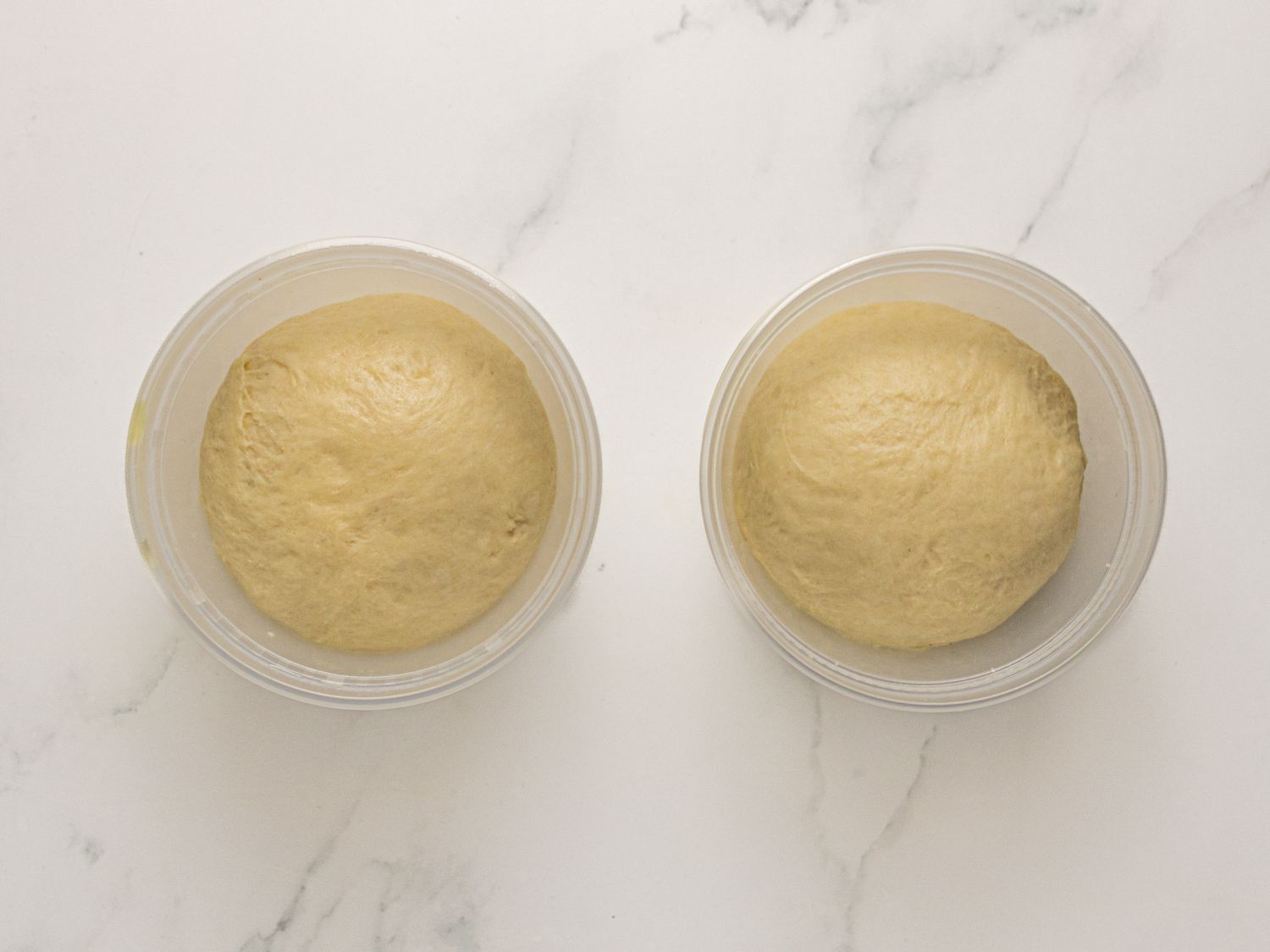 Two clear bowls with dough balls on a white surface likely prepped for baking