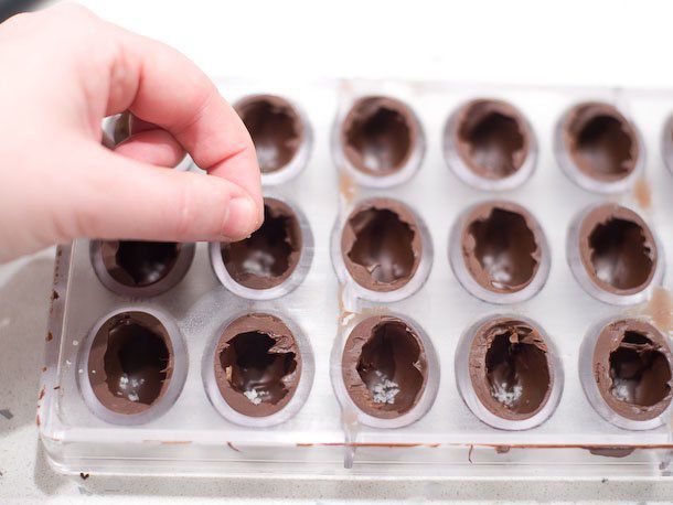 A hand sprinkling sea salt into bonbon molds already filled with a chocolate shell.