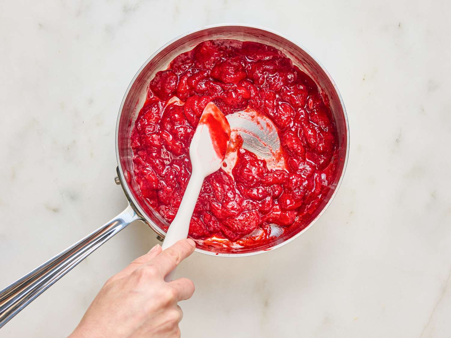 A saucepan with a strawberry mixture being stirred with a spatula by a hand