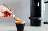 A hand holding a spoon in a cup of espresso next to the Nespresso VertuoPlus Coffee and Espresso Machine by De'Longhi
