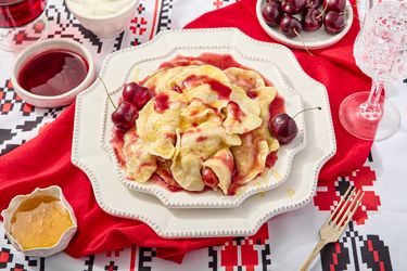 Cherry verenyky on decorative plates, on a printed surface, with a red napkin, and sides of cherry sauce, cream, and honey 