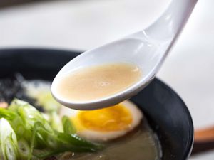 Closeup of a spoonful of chicken paitan broth. A bowl of ramen is visible in the blurred background.
