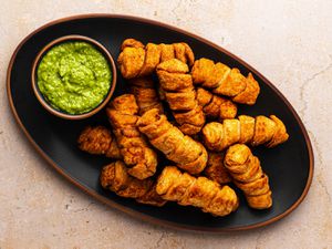 An oval black ceramic plate holding a pile of golden brown tequeÃ±os and a small bowl holding the avocado dipping sauce called guasacaca.