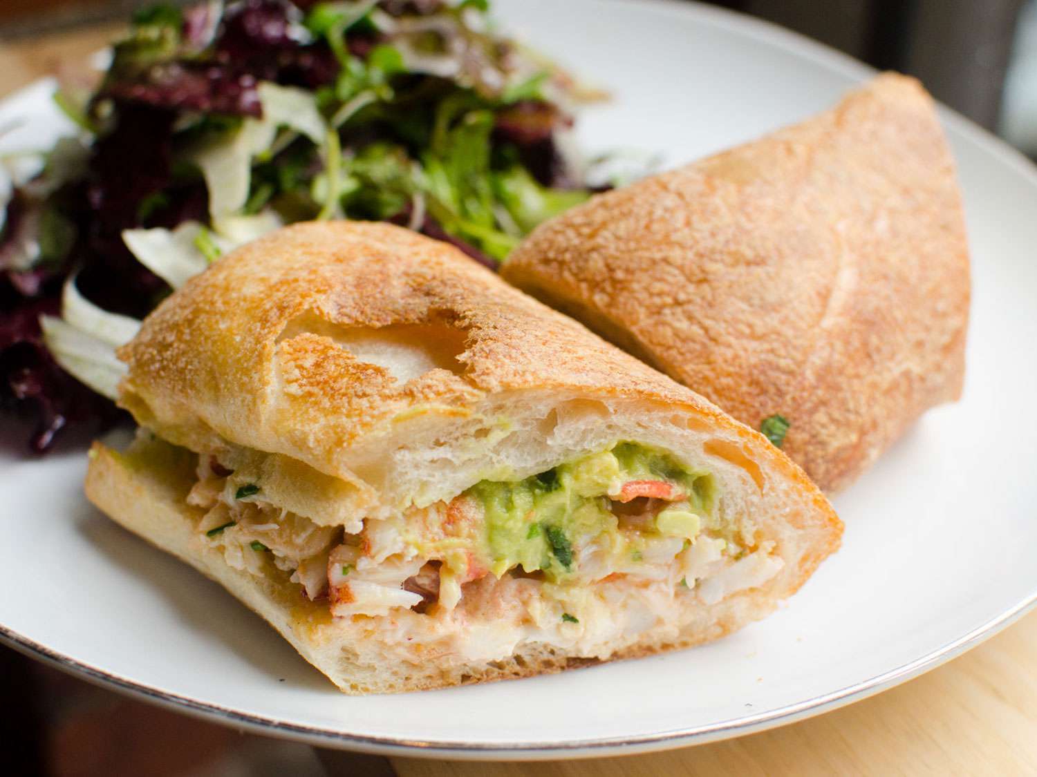 A lobster sandwich with avocado on ciabatta, served with a mesclun salad on the side.