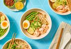 Bowls of curry laksa garnished with shrimp boiled egg cucumber and beans accompanied by lime and chili paste on a blue background