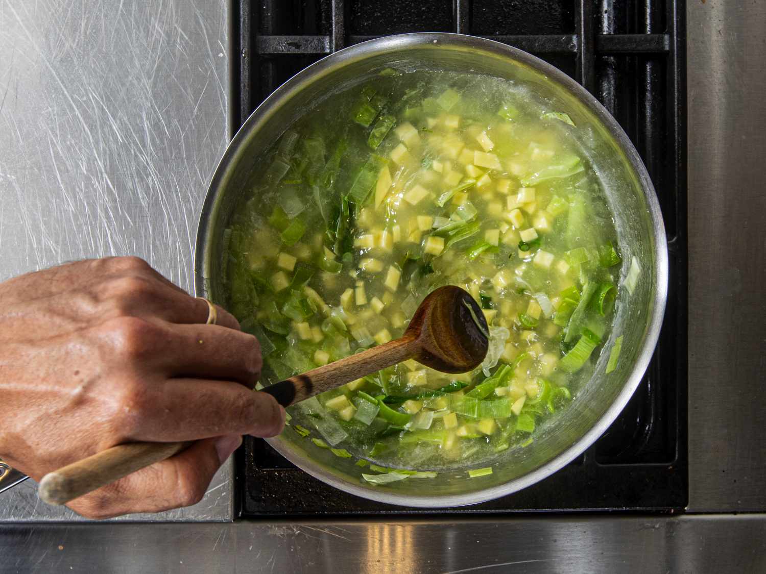 Overhead view of potato and water added to pot with garlic scapes and leeks
