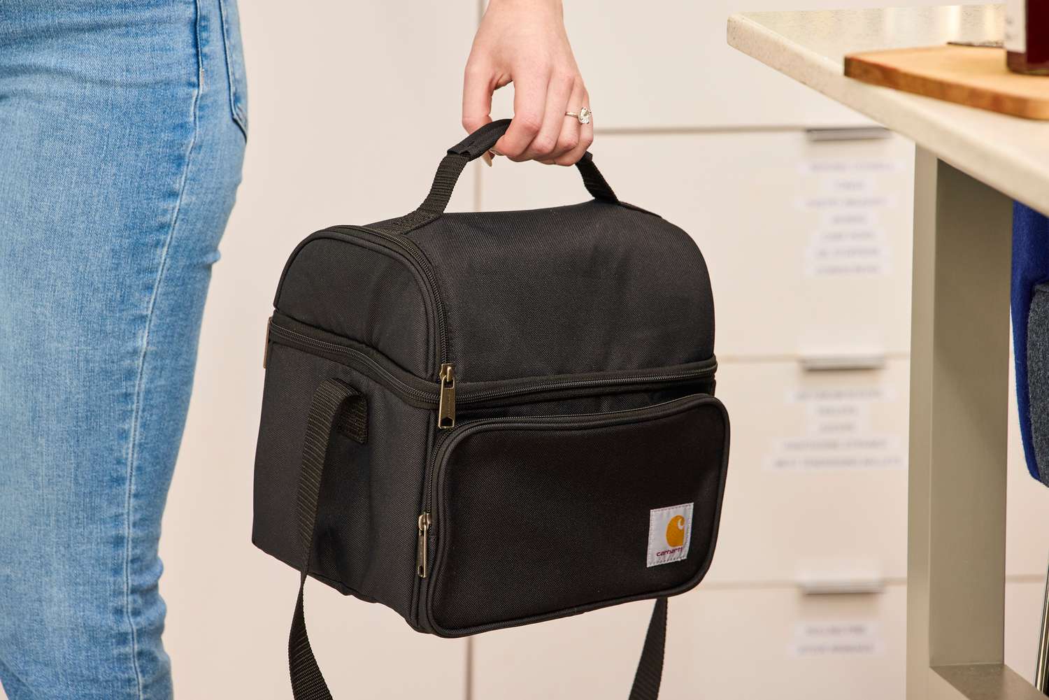 A hand holds the top handle of a black lunch box.