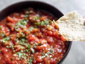Close-up of a chip topped with salsa. A bowl is in the background, slightly out of focus.