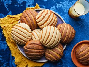 Overhead view of conchas on a plate with a glass of milk. Tablecloth is a velvety blue with a floral pattern and plate is resting on a bright yellow napking