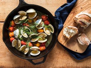 Broiled clams with tomatoes and tarragon in cast iron skillet beside wooden board with crusty bread