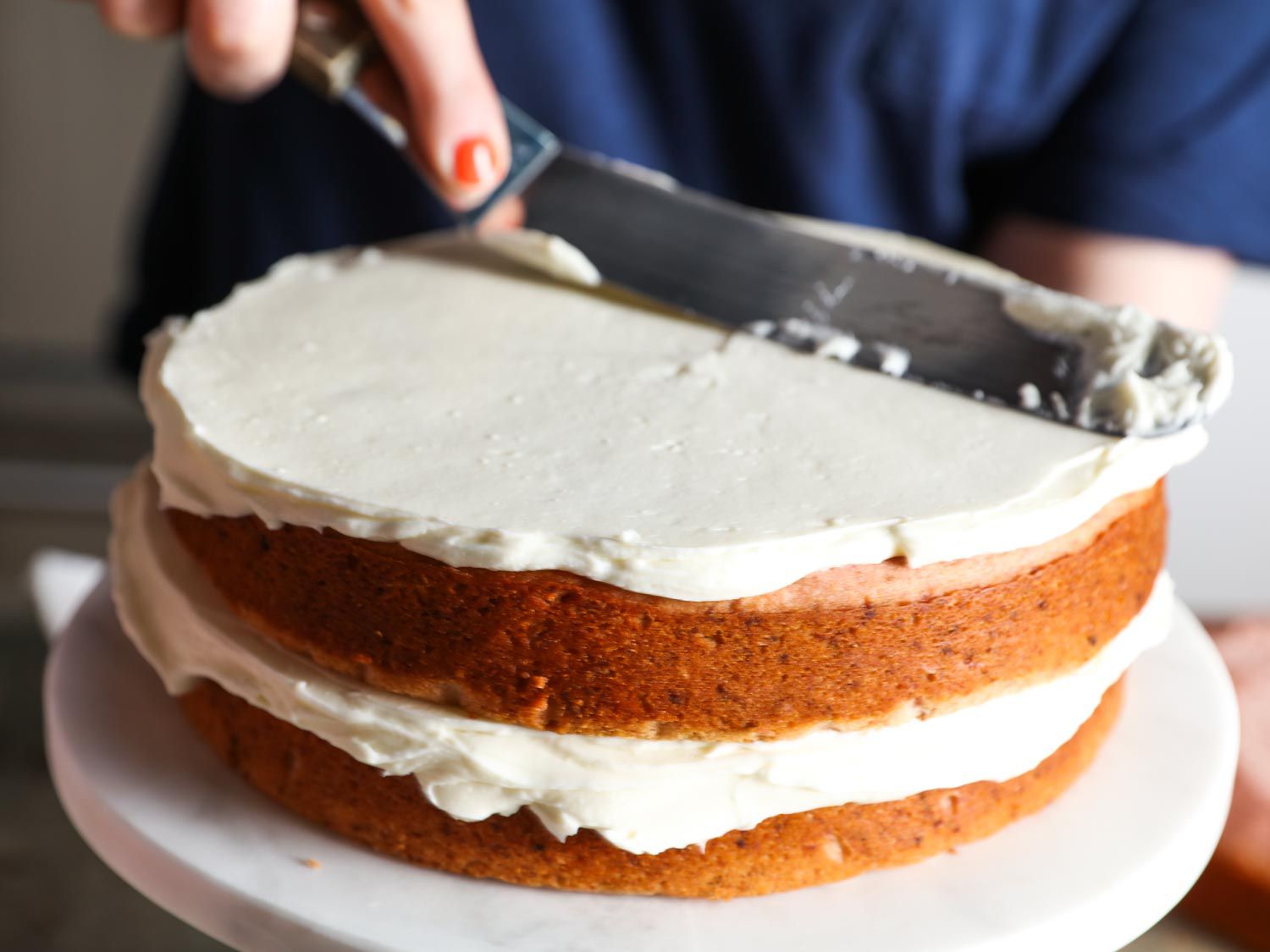 Using an offset spatula to spread cream cheese frosting over stacked layers of strawberry cake.