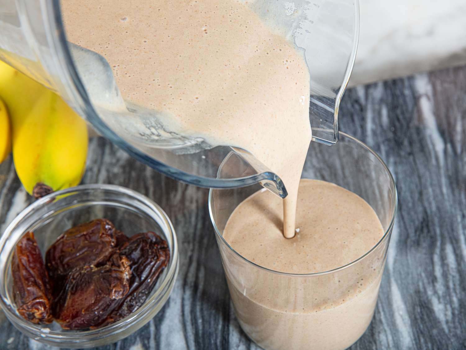 Pouring a banana and date smoothie from a blender into a glass bananas and dates visible on tabletop