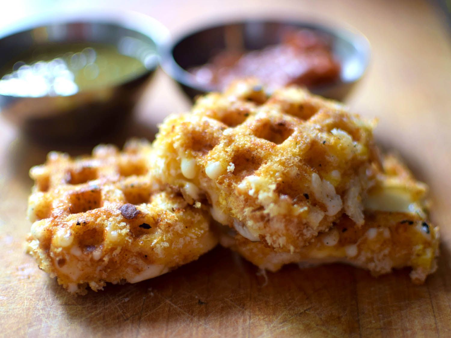 Closeup of waffle iron fried cheese, stacked on a cutting board. Dipping sauces are visible in the blurred background.