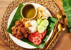 Plate of beef Rendang with rice, cucumbers, tomatoes and crisps on a banana leaf, with a woven placemat and gold cutlery