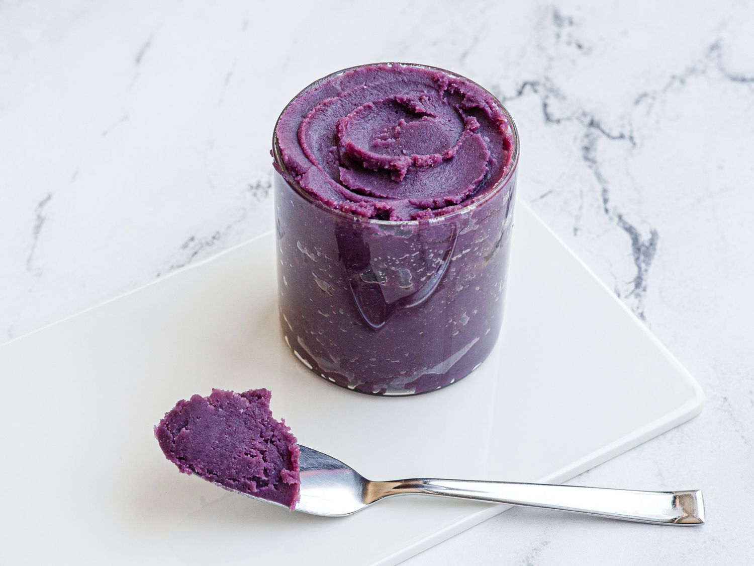 A glass jar of ube halaya placed on a white marble surface. A morsel of the stiff jam is perched on a spoon in front of the jar.