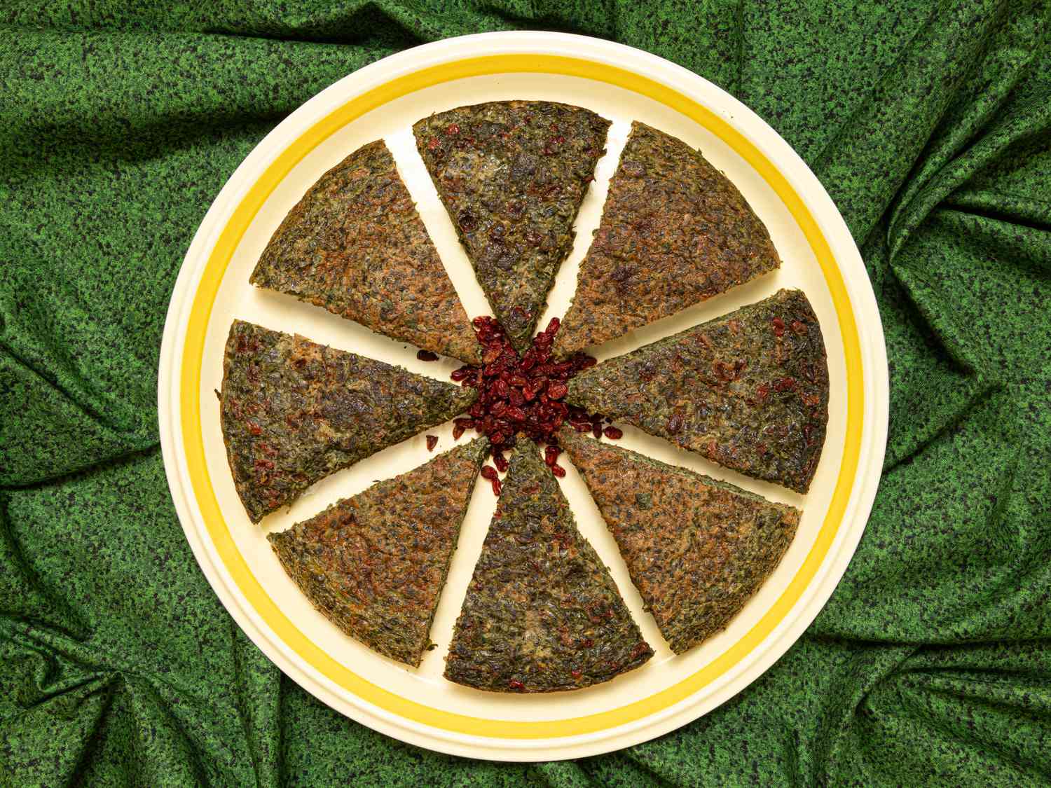 Sliced of herb kookoo arranged in a circular shape, with dried barberries in the center. Dish is on a green texture textile 