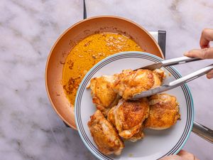 A plate with cooked chicken thighs being held above a copper pan