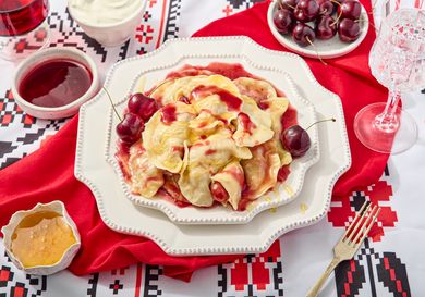 Cherry verenyky on decorative plates, on a printed surface, with a red napkin, and sides of cherry sauce, cream, and honey 