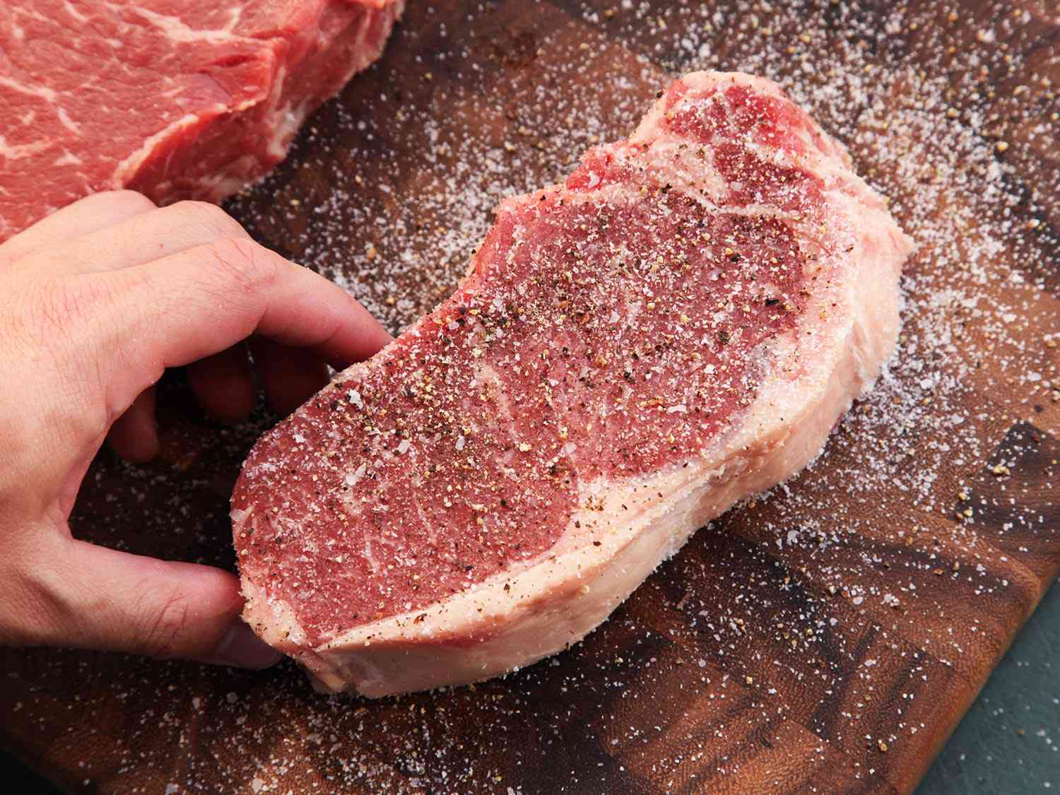 A raw steak on a cutting board being seasoned with salt and pepper. 