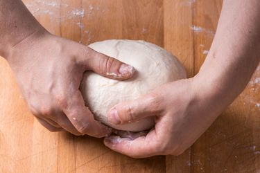 Two hands gently hold a smooth ball of dough on a wooden work surface.