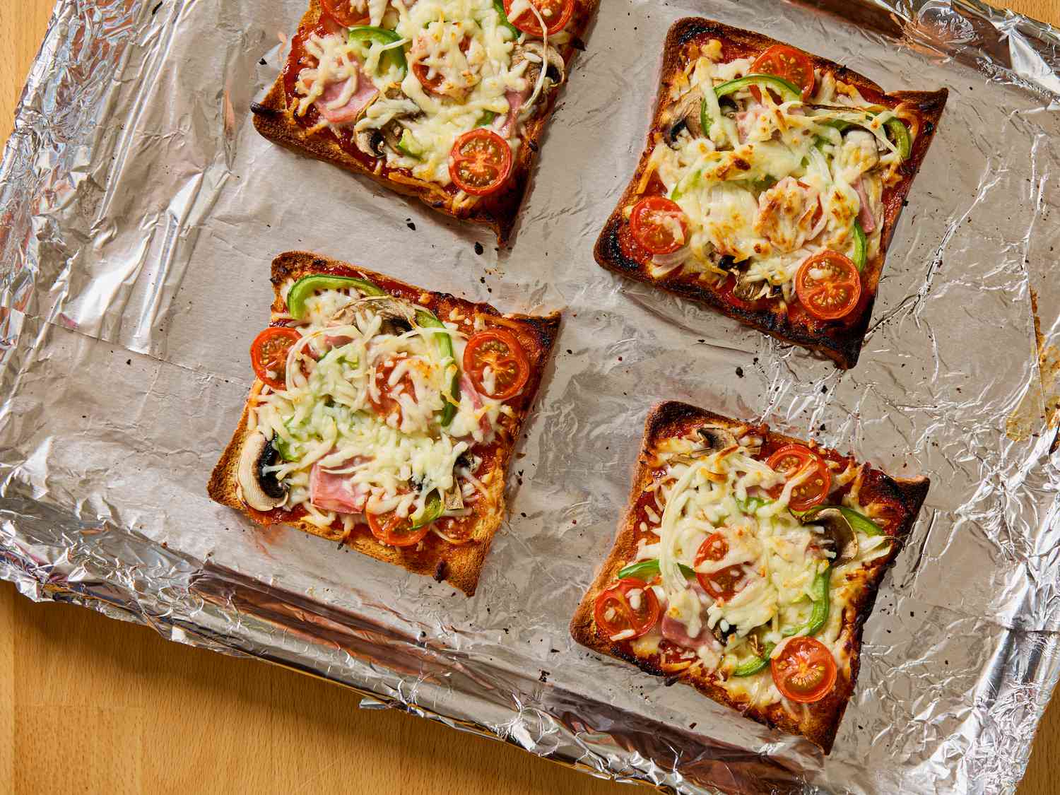 Four slices of pizza toast with vegetables and cheese, placed on a foil-lined baking tray