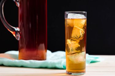 A tall glass filled with ice and cold-brewed iced tea, with a full glass pitcher of iced tea behind it.