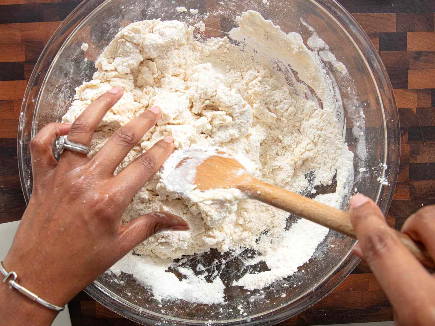 Hand and wooden spoon mixing together dough in glass bowl on wooden chopping block 