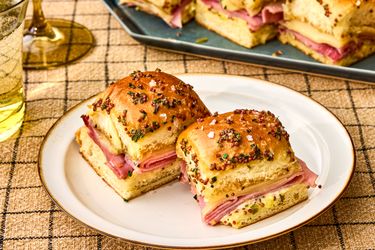 Ham and cheese sliders on a plate garnished with a poppy seed topping with additional sliders on a tray in the background