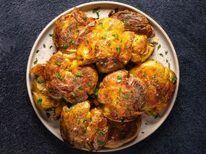 Golden brown crispy smashed potatoes on an oval white plate on a dark textured background.