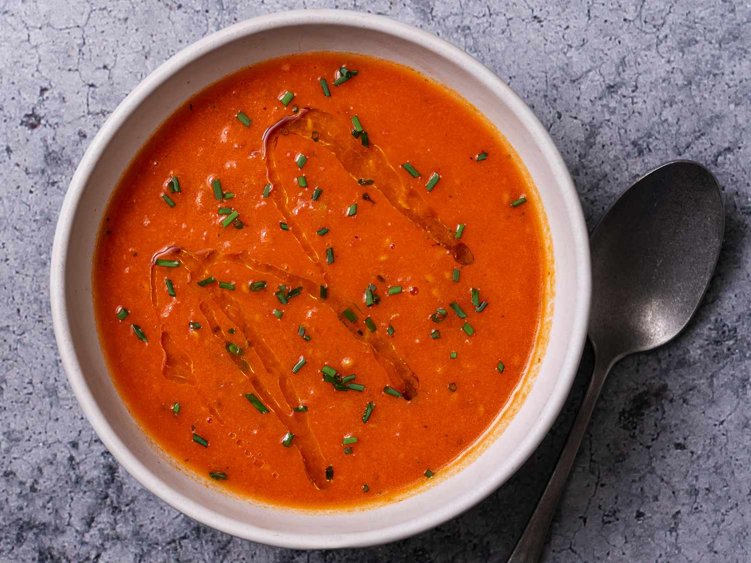 A bowl of creamy vegan tomato soup on a stone background with a spoon to the right of the bowl. The surface of the soup is drizzled with olive oil and sprinkled with chopped herbs.