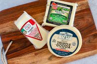 A wooden board holding three types of packaged cheese from Wisconsin Cheese Mart's Wisconsin Cheese of the Month Club, including Fontinella, Hatch Chile Cheddar and Truffle Gouda