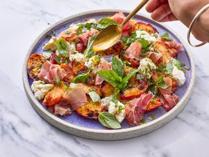 A hand drizzling dressing over a grilled peach salad with prosciutto and basil