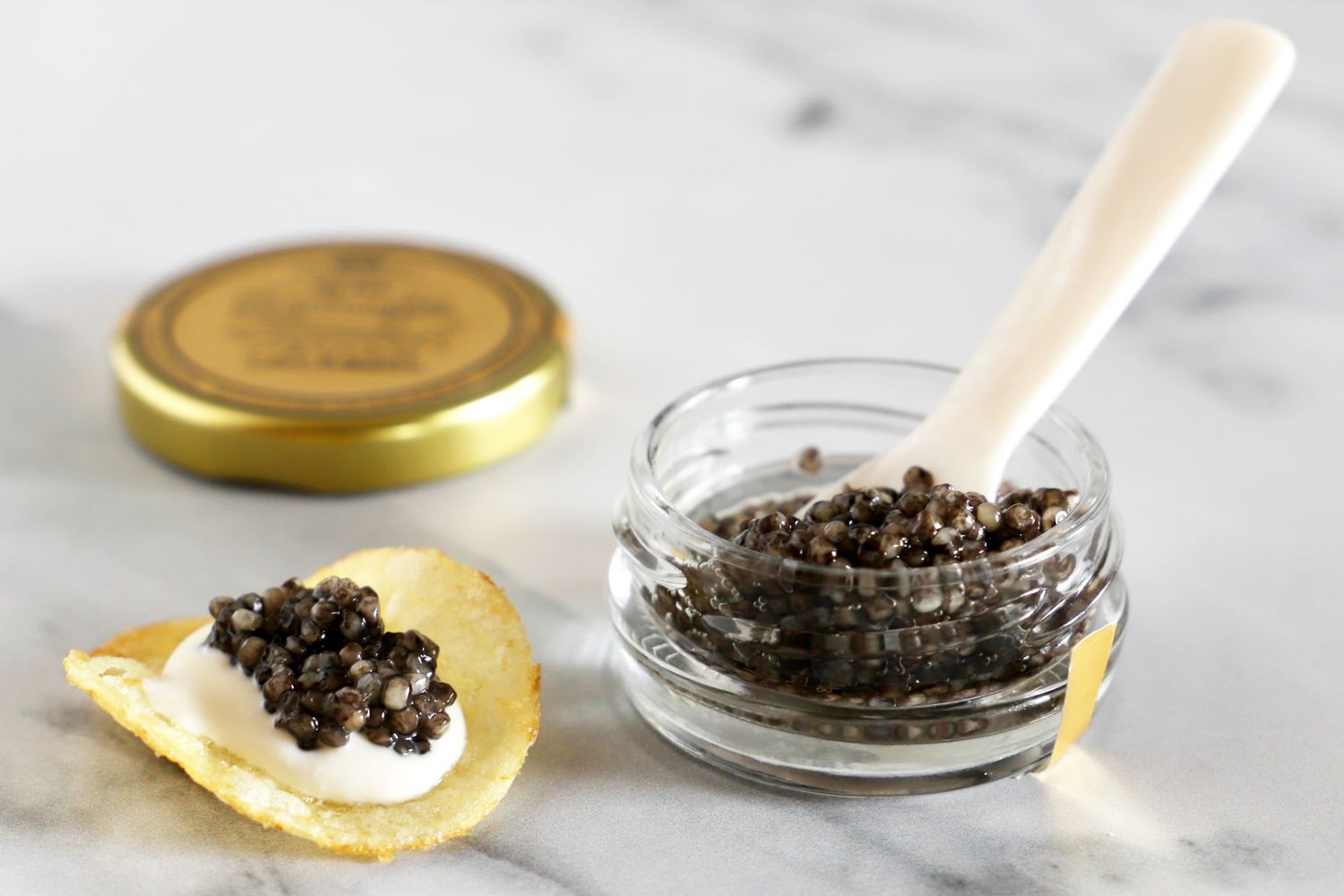 An open jar of Olma beluga hybrid caviar next to a potato chip topped with crème fraîche and a scoop of caviar