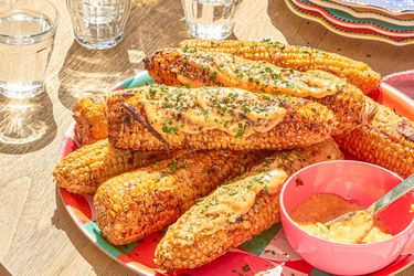 Plated grilled corn cobs topped with sauce and herbs alongside a small bowl of sauce