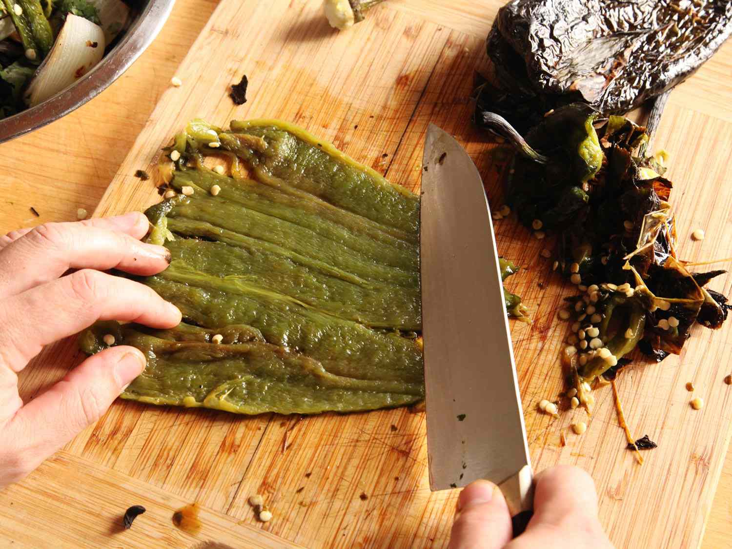 A peeled green chile is flattened and laid out on a cutting board. A santoku knife has scraped the seeds and ribs from the interior surface.