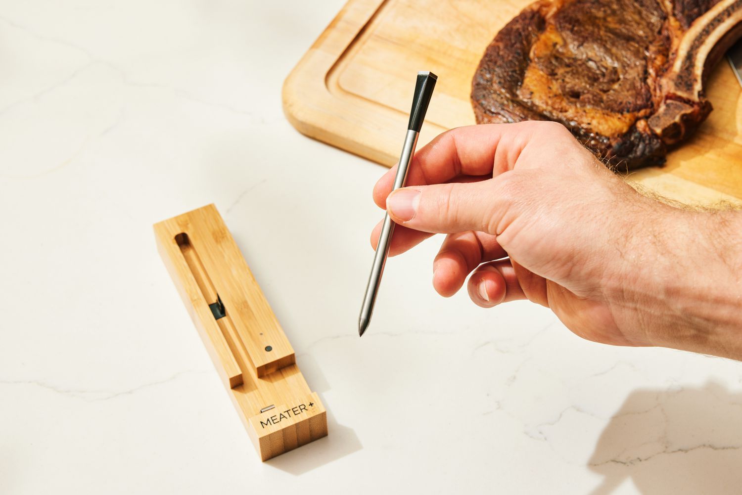 A hand holding the Meater Plus Smart Meat Thermometer with Bluetooth and the charger is displayed on top of a table below