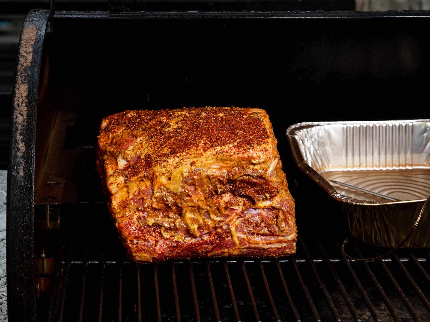 The seasoned pork shoulder is place on the grill grate of a smoker beside an aluminum tray containing water.