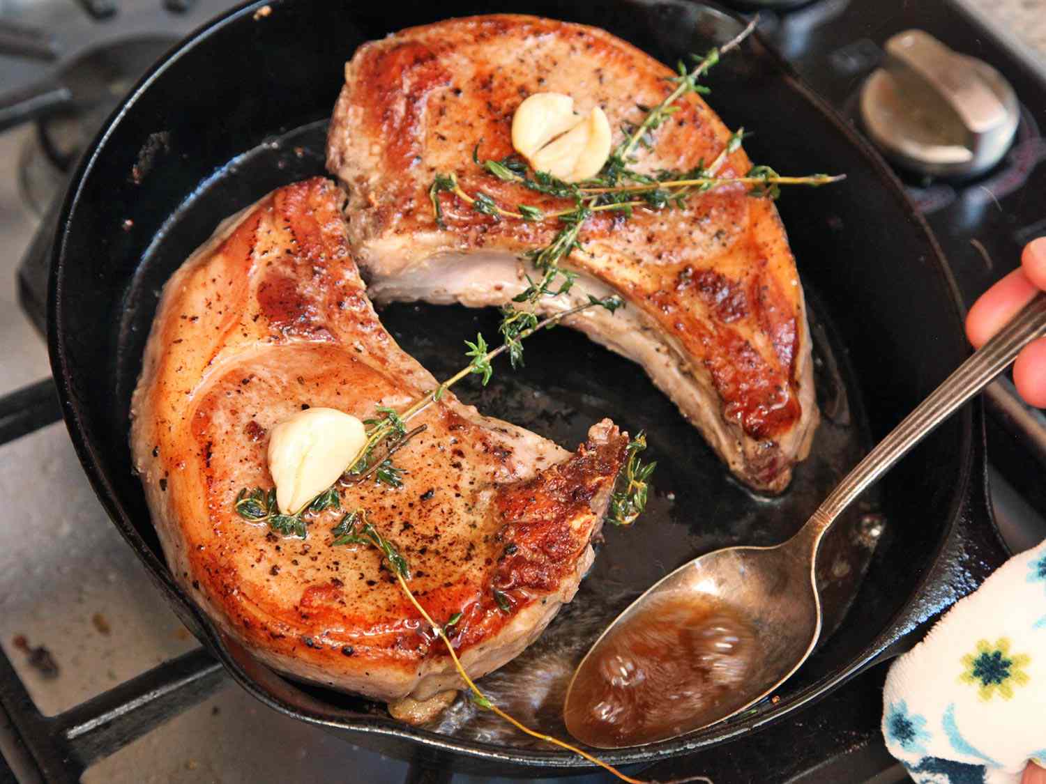 Two pork chops with butter, thyme, rosemary, and garlic cooking in a cast iron skillet.
