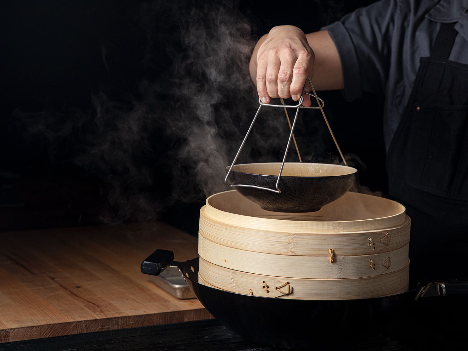 A hot dish steamer tong lifting a bowl out of a bamboo steamer set in a wok.