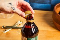 A hand with tattoos unscrews a gold cap from a dark glass bottle, next to measuring spoons and scattered grains