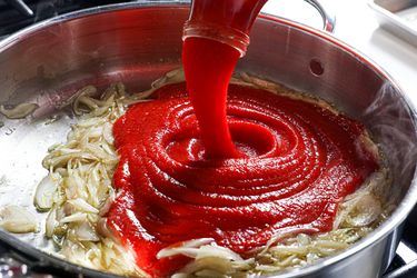 Tomato puree being poured into a pan with sautéed onions on a stovetop surface