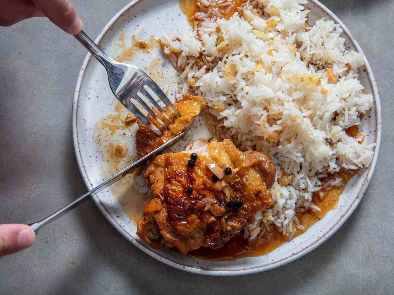 Overhead view of knife and fork cutting into chicken thigh adobo on a plate with garlic fried rice