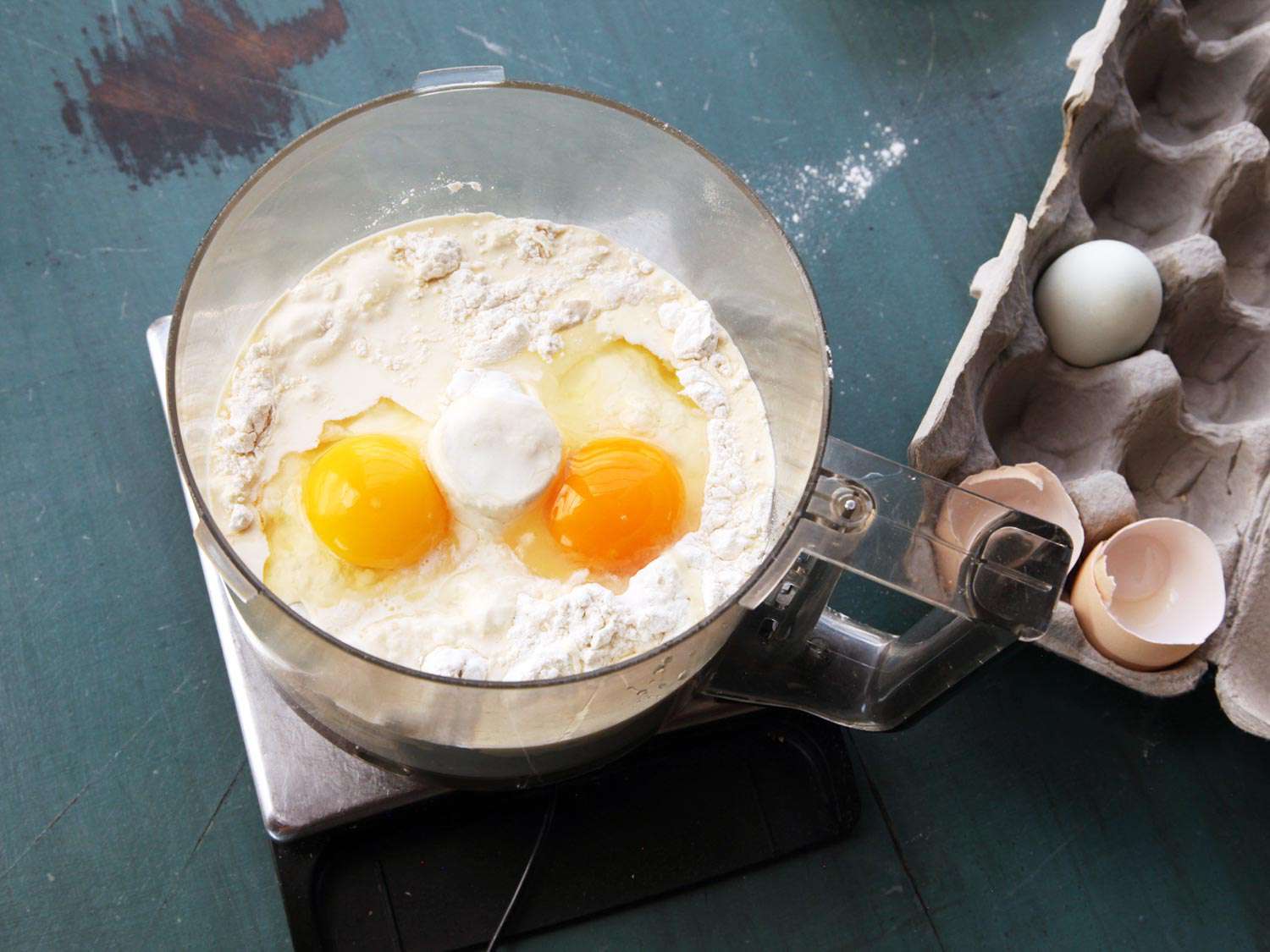 Flour and eggs are added to the bowl of a food processor, which has been placed on a digital scale.